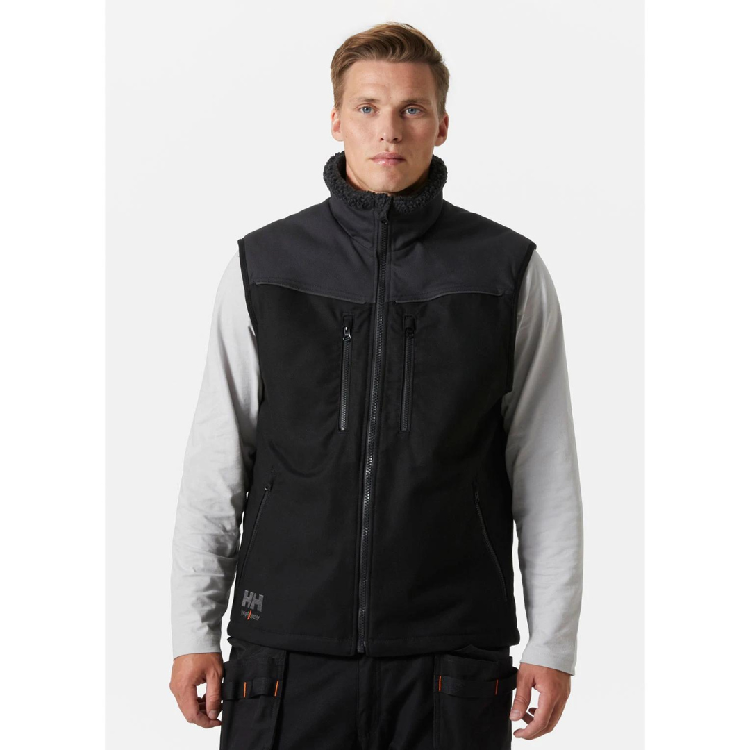 VESTE DE TRAVAIL DOUBLÉE SANS MANCHE HELLY HANSEN WORKWEAR OXFORD couleur black/ebony porté par un homme vu de face veste entièrement noire on voit les deux textures des matières, les fermetures éclaires des 4 poches extérieures ainsi que le logo hh workwear au bas droit de la veste