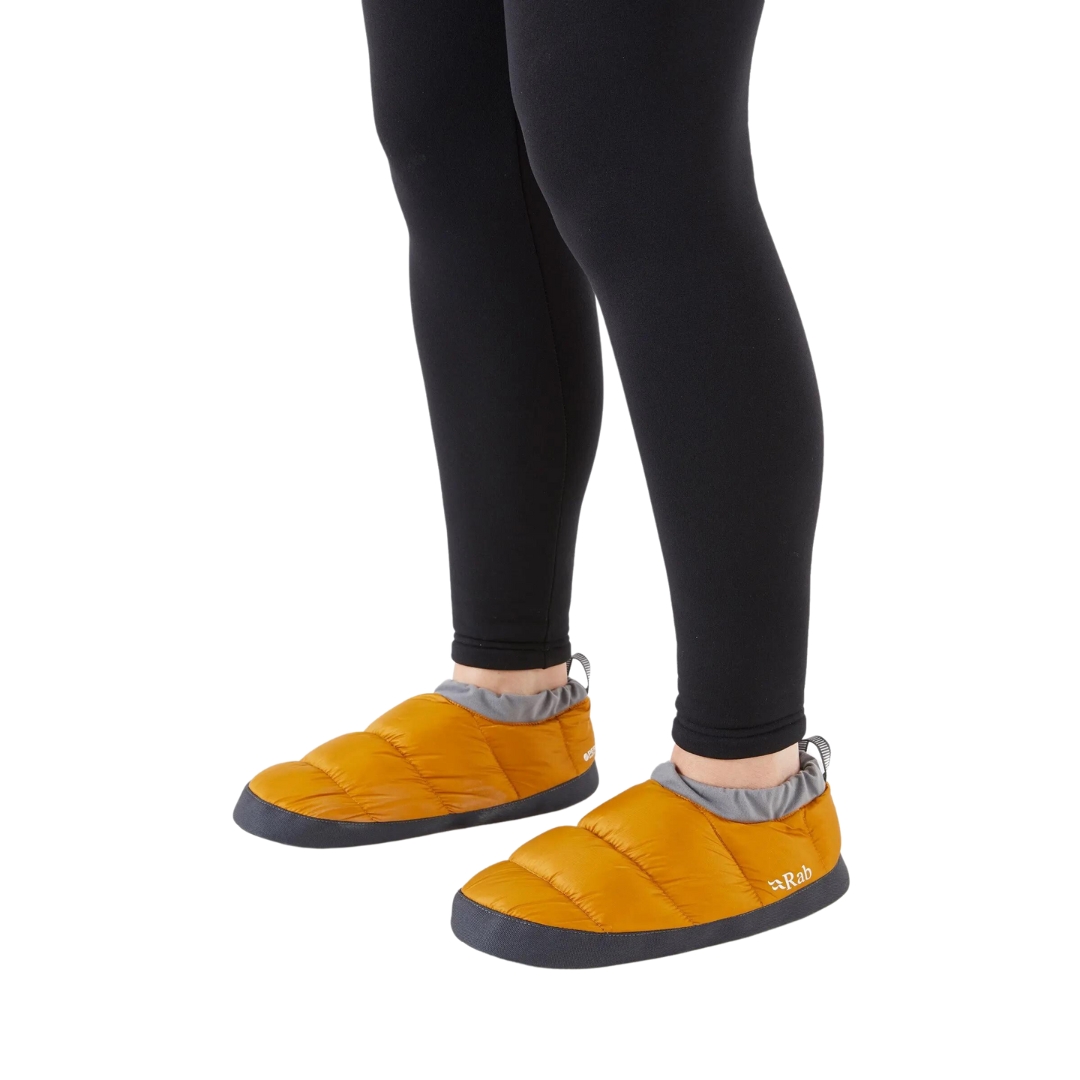 CHAUSSON DE BIVOUAC RAB DOWN HUT UNISEXE ADULTE COULEUR DARK BUTTERNUT VU DES CHAUSSON BAS DE COULEUR JAUNE OCRE PORTÉ PAR UNE FEMME VUE DES GENOUX AUX PIEDS DE PROFIL AVANT GAUCHE