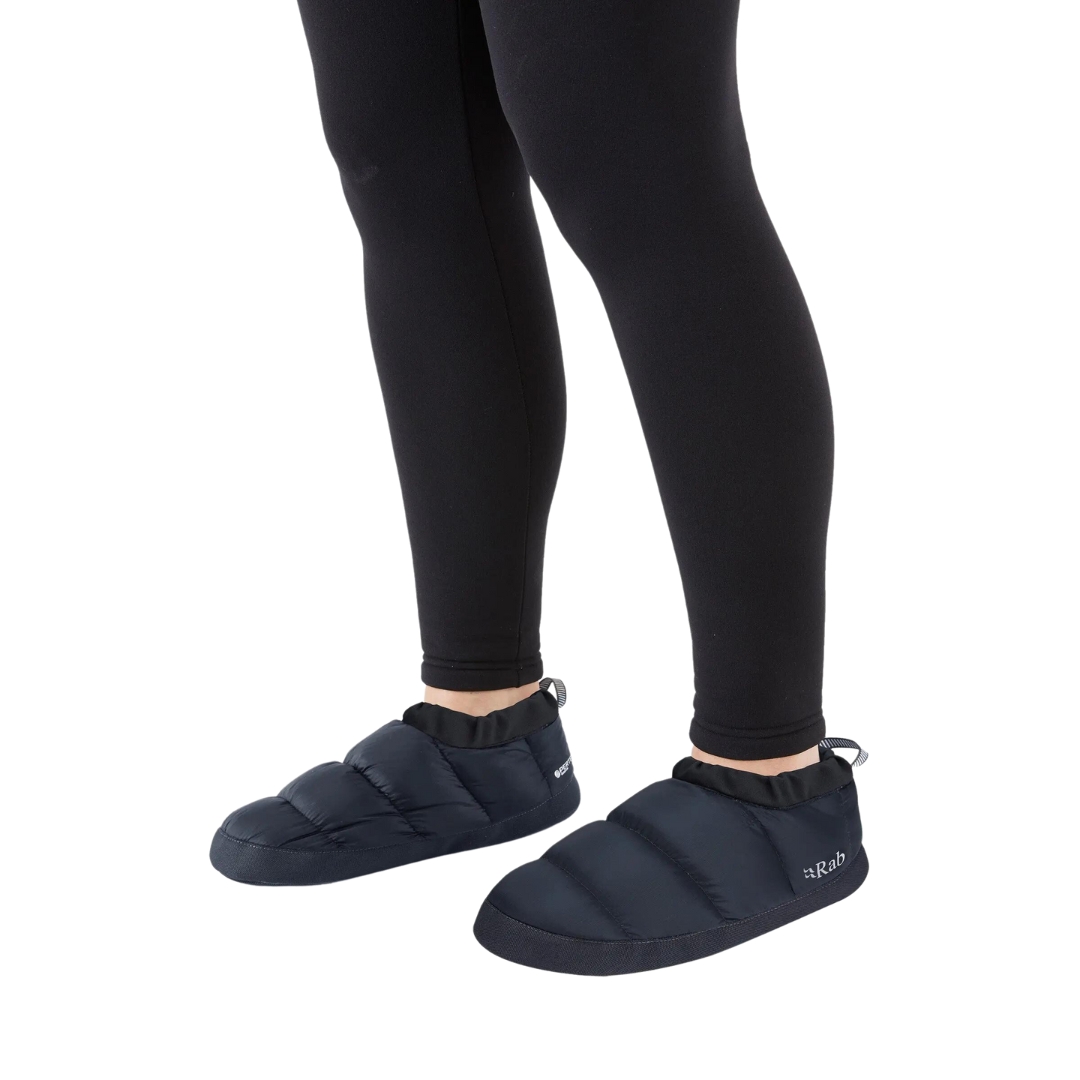 CHAUSSON DE BIVOUAC RAB DOWN HUT UNISEXE ADULTE COULEUR BELUGA VU DES CHAUSSON BAS DE COULEUR NOIR PORTÉ PAR UNE FEMME VUE DES GENOUX AUX PIEDS DE PROFIL AVANT GAUCHE