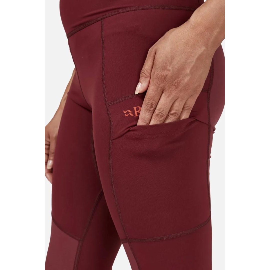 LEGGING D'ENTRAINEMENT RAB HORIZON POUR FEMME couleur Deep Heather porté par une femme qui glisse une main dans la poche de la cuisse gauche avec le logo et nom RAB imprimé en rouge pale sur le rouge brique du pantalon
