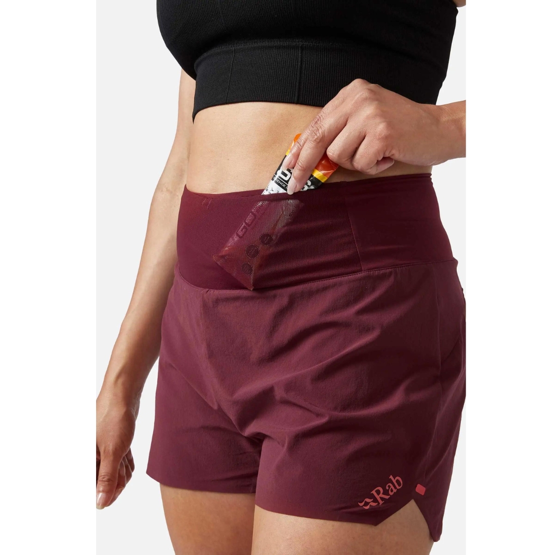 SHORT D'ENTRAINEMENT RAB TALUS TRAIL LIGHT 5'' POUR FEMME porté par une femme qui range une barre énegétique dans la poche `afilet a l'avant