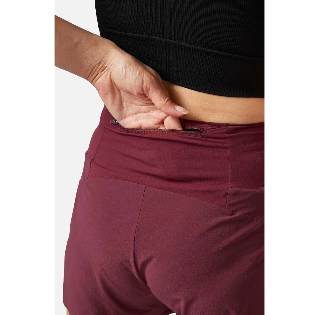 SHORT D'ENTRAINEMENT RAB TALUS TRAIL LIGHT 5'' POUR FEMME porté par une femme qui montre l'ouverture de la poche de taille arrière avec sa main