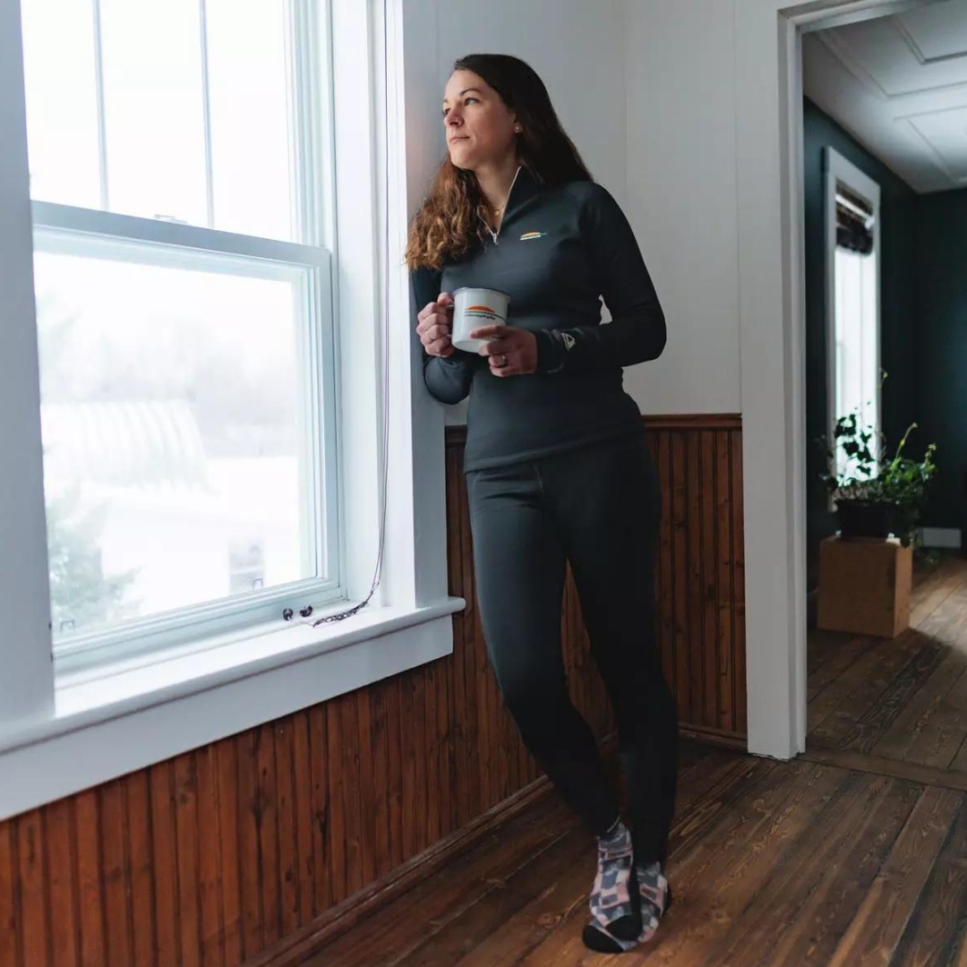 LEGGING COUCHE DE BASE CHLOROPHYLLE CHL ULTRA POUR FEMME couleur noir pur vu du legging noir avec band élastique a la taile brune porté par une femme vu e de la tête aux pieds dans un chalet avec une tasse regardant par la fenêtre