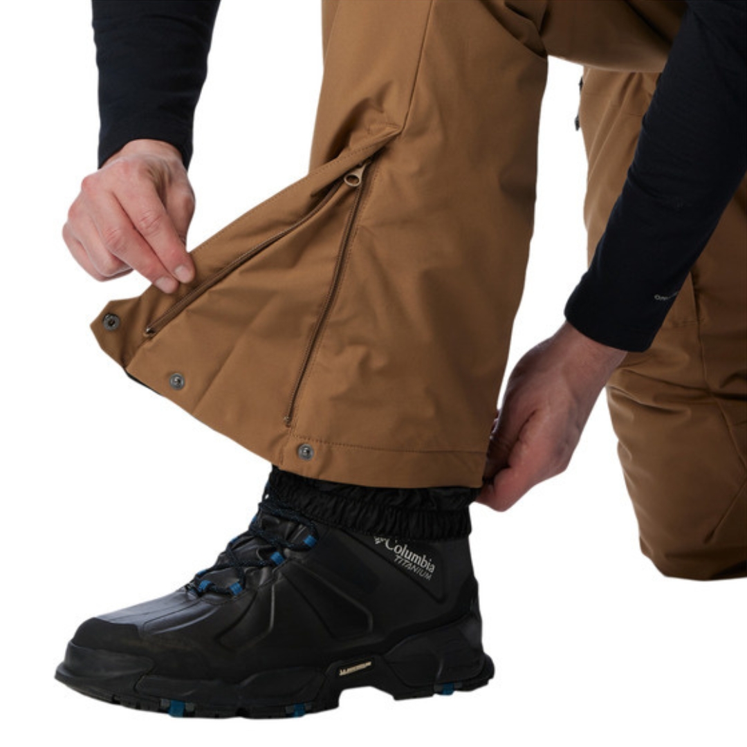PANTALON DE NEIGE COLUMBIA KICK TURN III POUR HOMME couleur 257-DELTA vu du pantalon brun sable porté par un homme vu en gros plan sur le bas du pantalon l'homme montre le souffle ajustable à la cheville