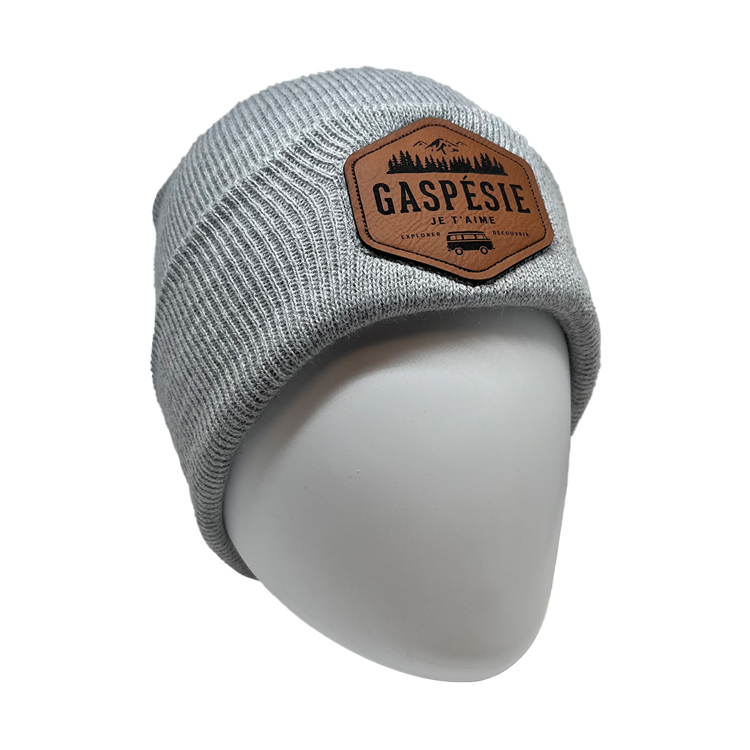 Un bonnet tricoté gris avec un patch en cuir sur le devant. Le patch comporte une illustration de montagnes et d'arbres, ainsi que le texte « Gaspésie Je T'aime » et un texte plus petit en dessous. Le bonnet est présenté sur une tête de mannequin blanche unie.