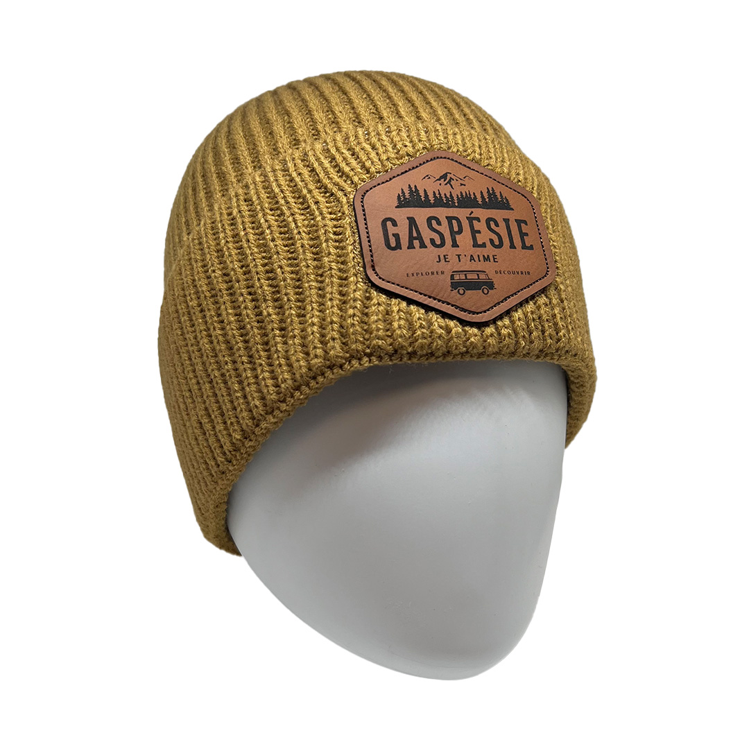 Un bonnet en tricot jaune moutarde avec un patch marron sur lequel figure le texte « Gaspésie Je t'aime » et un motif de montagnes, d'arbres et d'une roulotte. Le bonnet est présenté sur une tête de mannequin blanche.