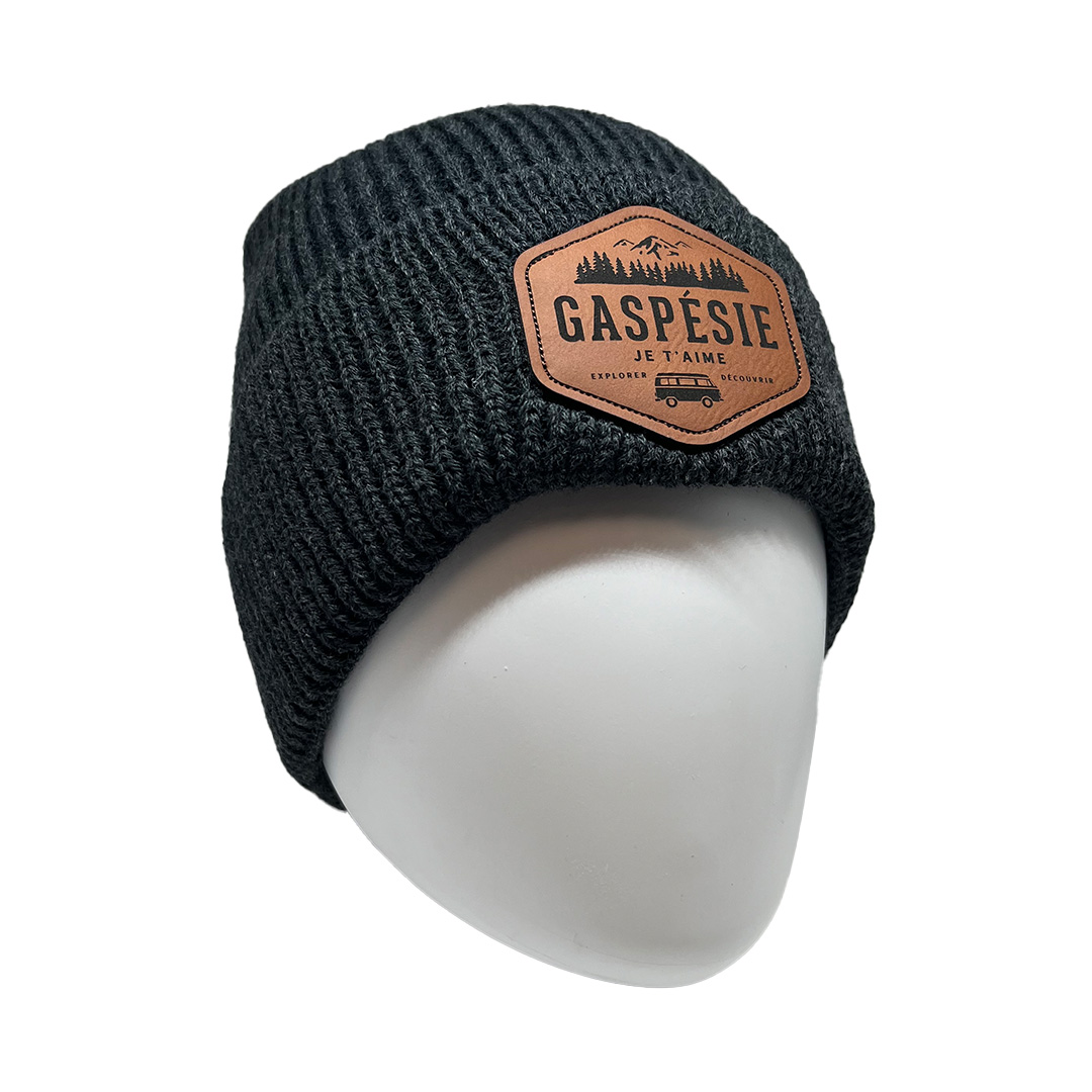 Un bonnet en tricot noir avec un patch en cuir beige sur le devant, portant l'inscription « GASPÉSIE JE T'AIME » ainsi qu'une illustration d'une chaîne de montagnes et d'arbres. Le bonnet est présenté sur une tête de mannequin blanche.