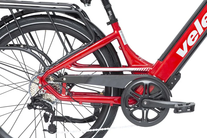Gros plan de la partie arrière d'un vélo rouge, montrant la chaîne, les vitesses, les pédales et une partie du cadre avec la lettre « v » visible. Il comprend un porte-vélo arrière et une béquille latérale. Le vélo est doté d'un design à enjambement.