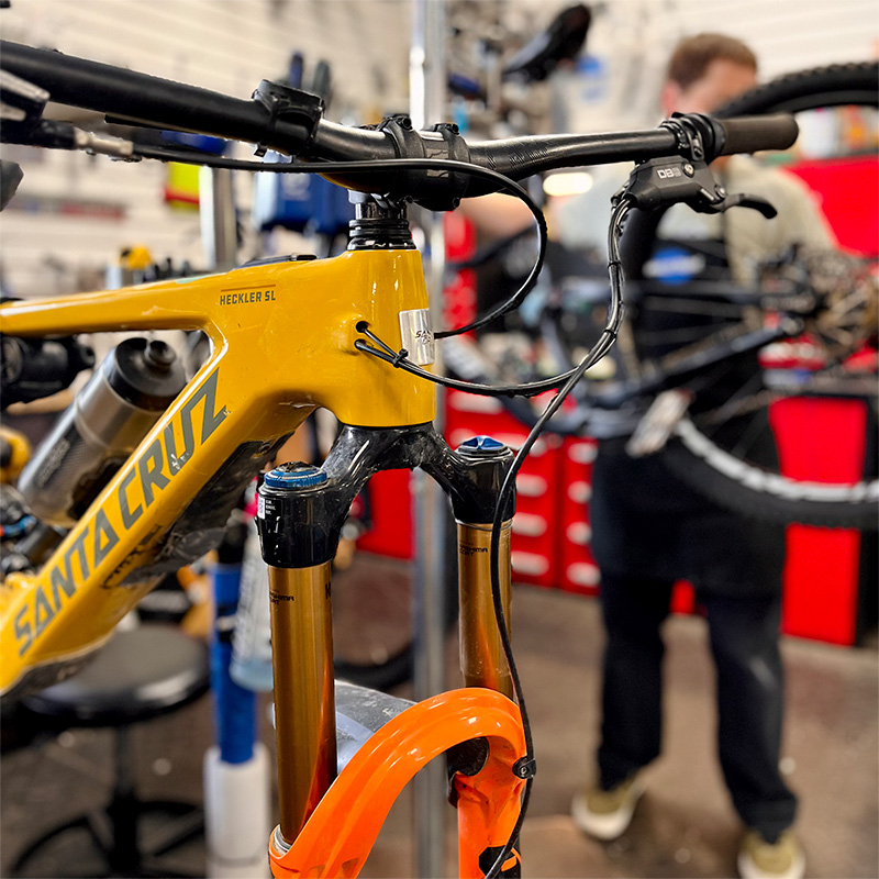 Gros plan sur l'avant d'un vélo de montagne Santa Cruz jaune dans un atelier de sport, avec un mécanicien à l'arrière-plan tenant un autre vélo. L'image met en évidence le guidon et la suspension avant.