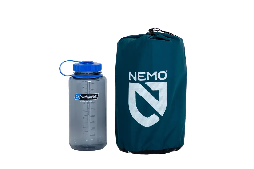 Une bouteille d'eau Nalgene bleue avec un bouchon bleu se trouve à côté d'un sac de couchage Nemo vert emballé dans un sac de rangement, tous deux sur un fond blanc.