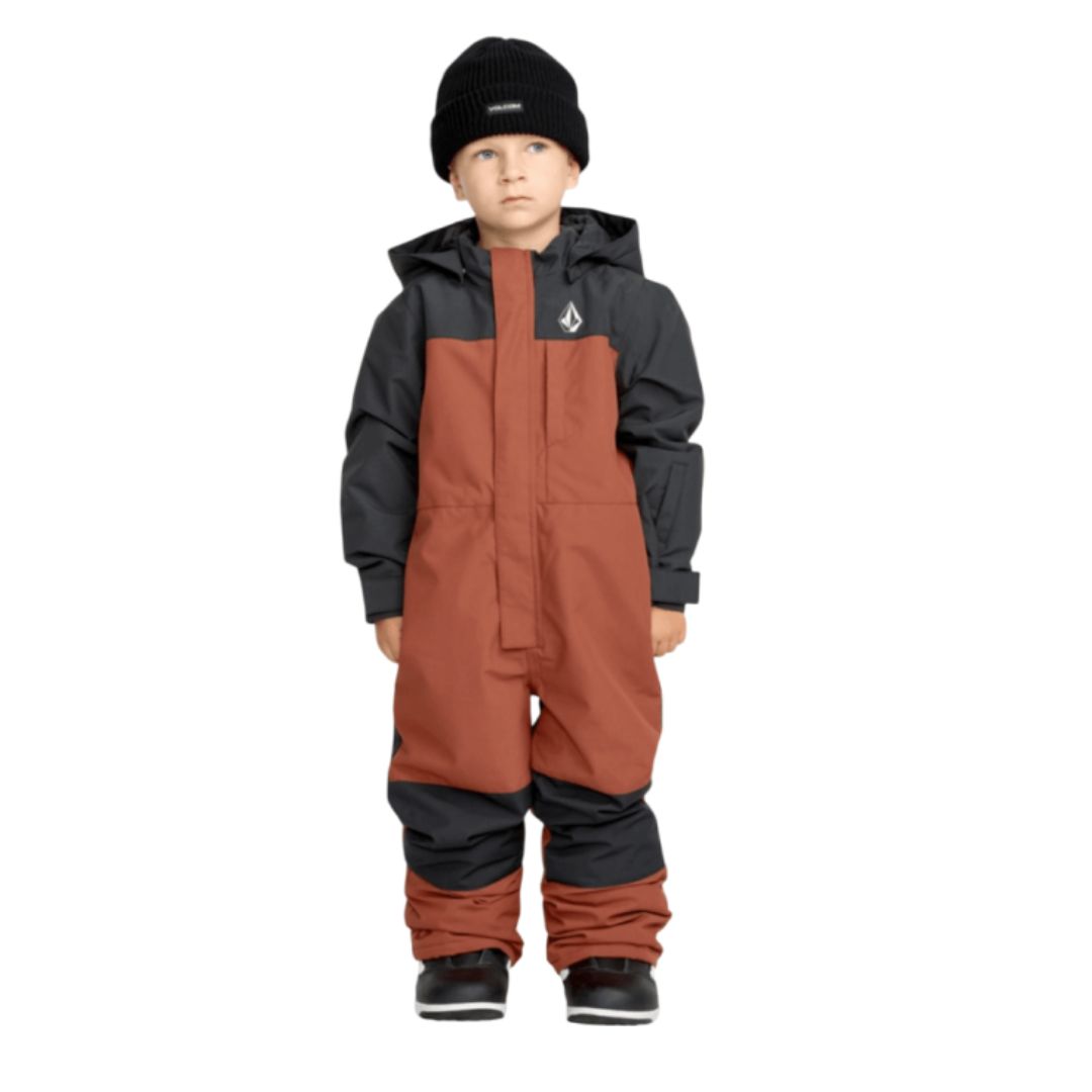 Un jeune enfant portant l'habit de neige LITTLE FLURRY ONE PIECE orange rouille et gris foncé avec une capuche, un bonnet noir et des bottes noires, se tient sur un fond blanc.