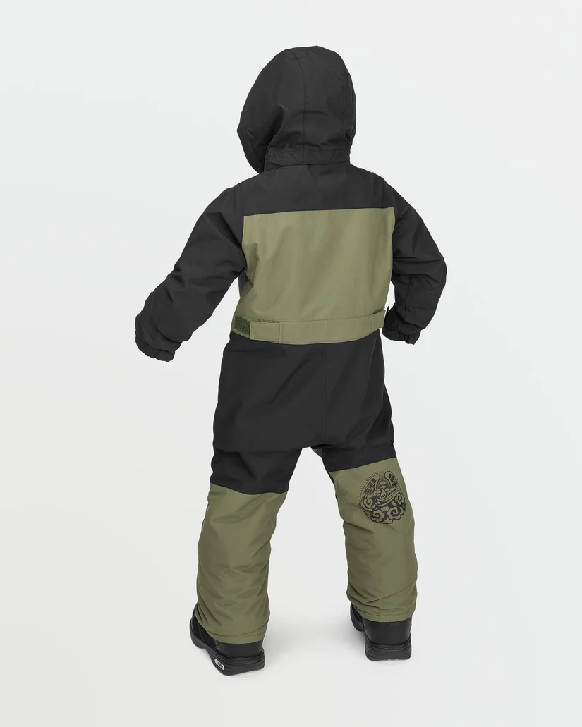 Un enfant porte une combinaison de neige à capuche vert foncé et noire et se tient dos à la caméra. La combinaison de neige couvre tout le corps, y compris la capuche et les jambes, avec des bottes noires qui dépassent du bas. La combinaison de neige a un motif circulaire à l'arrière de la jambe droite.