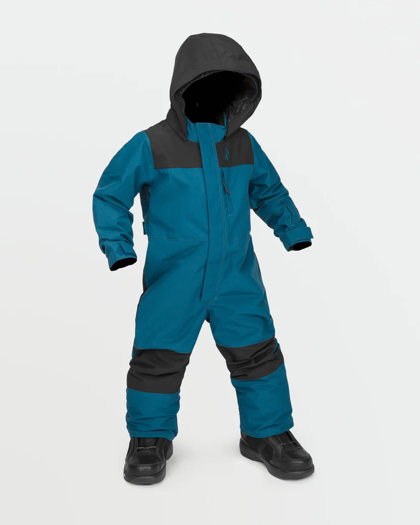 Un enfant porte une combinaison de neige bleu sarcelle et noire avec une capuche, debout sur un fond blanc uni. La combinaison de neige a des sections noires sur les épaules, les genoux et le bas des jambes, avec une fermeture éclair sur le devant et des bottes noires. Le visage n'est pas visible.