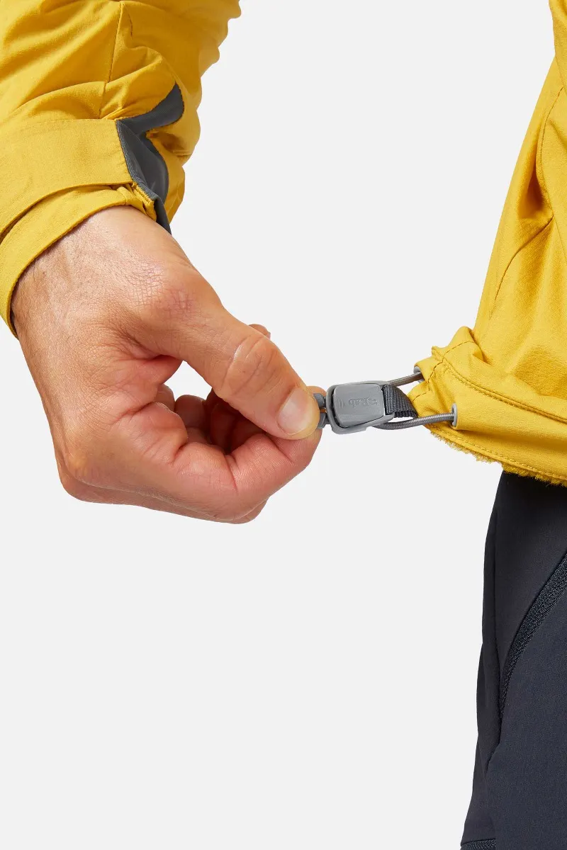 Une main ajuste le cordon d'une veste jaune, tirant sur un bouton noir pour le resserrer ou le desserrer autour de la taille. Le pouce et l'index de la personne maintiennent fermement le bouton. Le tissu de la veste a une apparence texturée et légère.