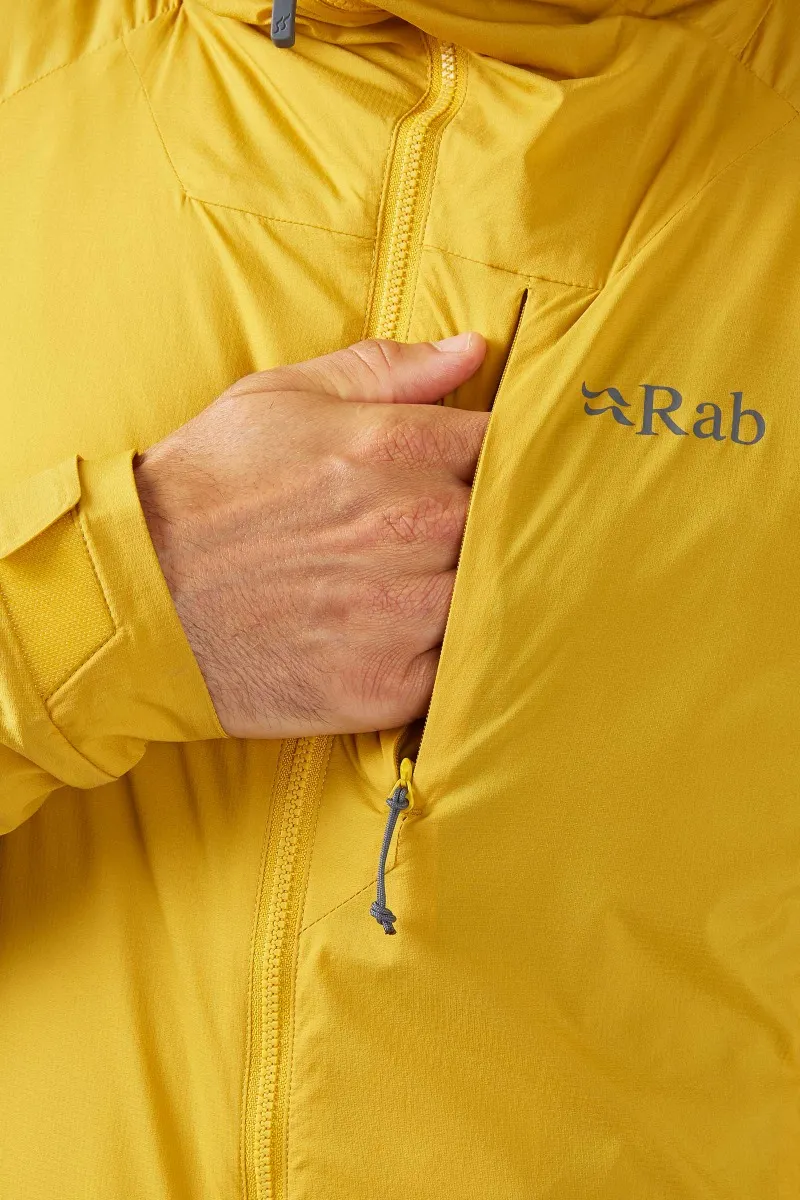 Une personne portant une veste Rab jaune ferme une poche de poitrine avec une main partiellement à l'intérieur. La veste présente une couture subtile et un petit logo sur le côté droit.