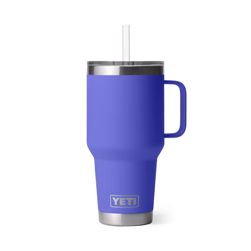 Un gobelet de voyage YETI bleu avec une poignée, un bord et une base en acier inoxydable et une paille en plastique transparent, debout sur un fond blanc.