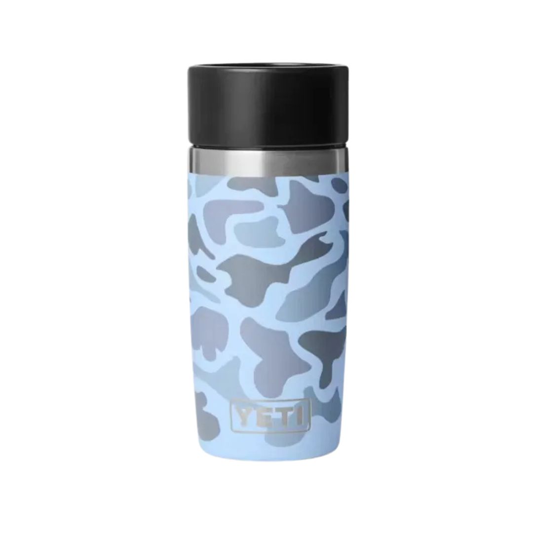 Une bouteille de voyage RAMBLER 12 oz CAMO, arborant un motif camouflage bleu sur le corps et dotée d'un bouchon noir, avec la marque « YETI » imprimée près du fond, est représentée sur un fond blanc uni.